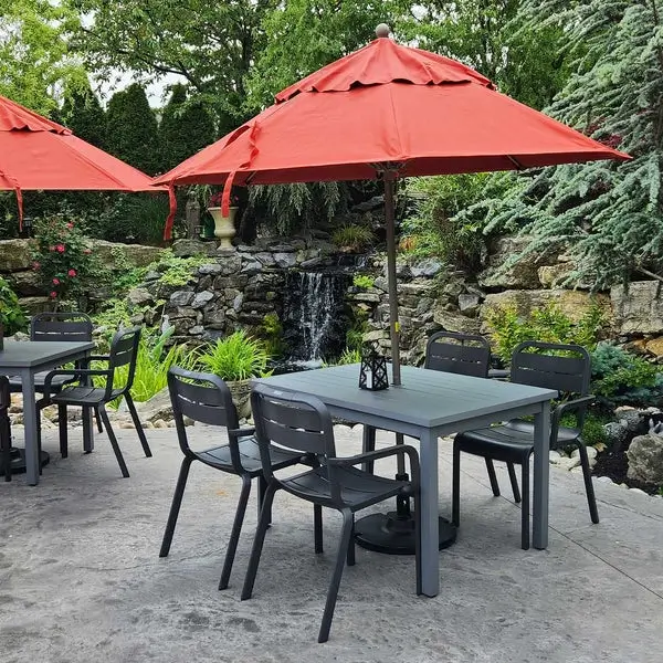 Commercial outdoor dining patio featuring modern Sigma dining tables, charcoal Cannes armchairs, and Terra Cotta Windmaster umbrellas beside a landscaped waterfall.